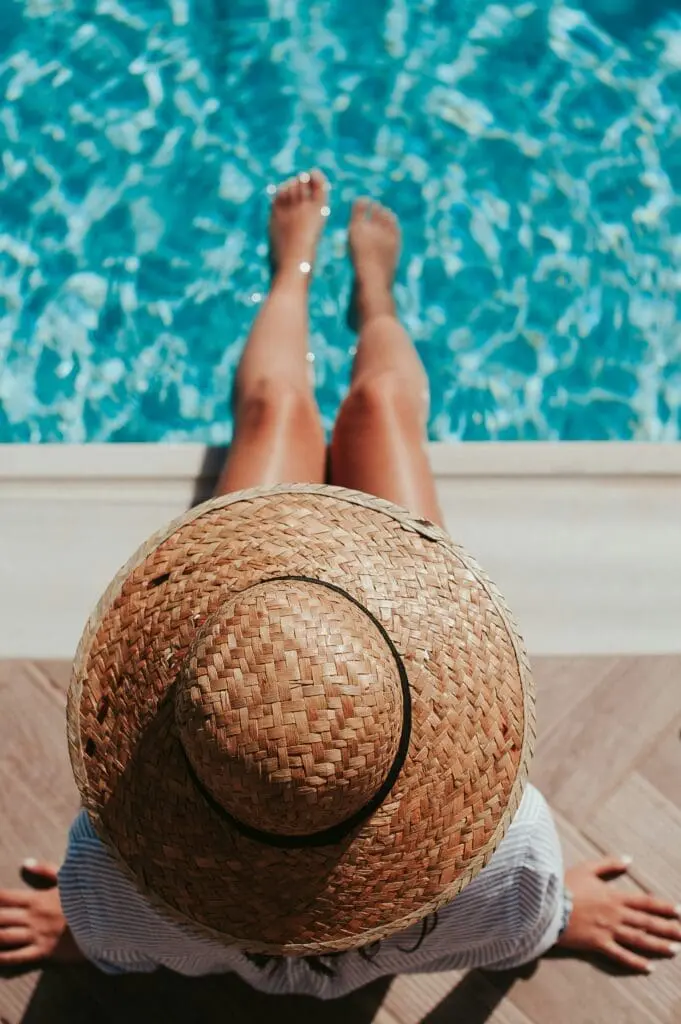 Woman wearing a straw hat sitting by a poolside with legs stretched out over turquoise water on a sunny day