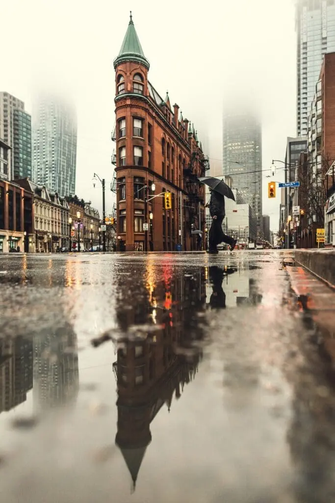 Person with umbrella crossing a rainy street in front of the Flatiron-style red brick Gooderham Building in downtown Toronto with reflections in puddles and foggy skyscrapers in the background