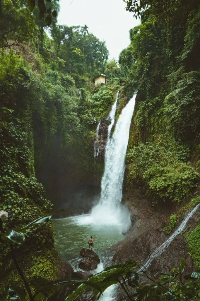 Man standing on a rock at the base of a tall jungle waterfall surrounded by lush green rainforest