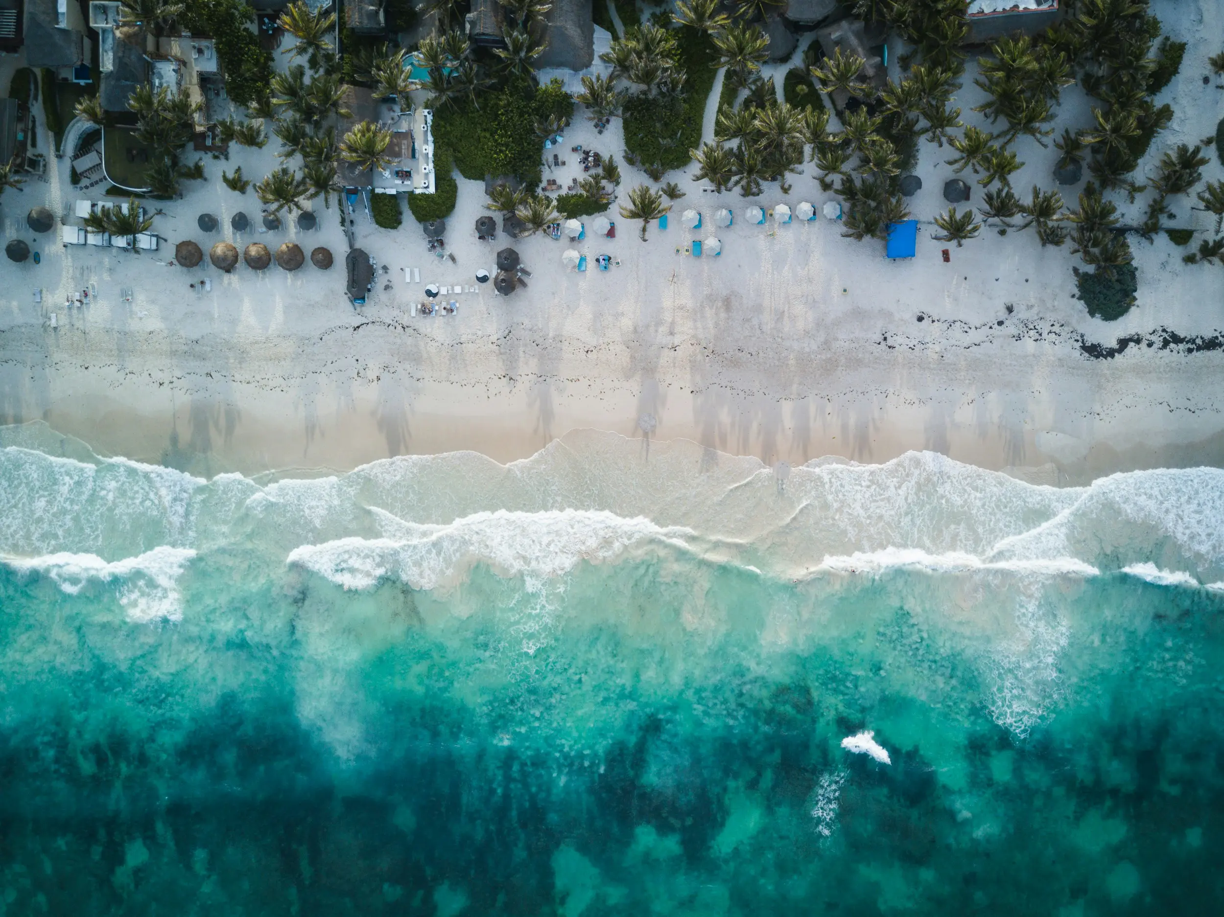 Aerial view of turquoise waves washing onto white sandy beach lined with palm trees and umbrellas in tropical resort